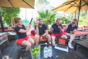 Cinq personnes assises sur des bancs rouges sous des parasols jaunes sur une terrasse en bois surplombant un lac, tenant des microphones et des téléphones lors d'un événement de team building près de Bordeaux.