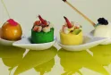 Quatre amuse-bouches gourmands présentés sur des assiettes en forme de cuillère blanche avec reflets sur une surface jaune-vert. De gauche à droite : une sphère dorée garnie de fleur rose, un gâteau rond vert surmonté de fruits de mer, un gâteau rond jaun