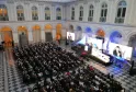 Large assemblée de conférenciers assis dans le Palais de la Bourse de Bordeaux, avec plusieurs écrans vidéo affichant des présentateurs et des présentations sur scène, sous un plafond à caissons orné et des fenêtres en arche.