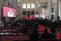 Grand hall de conférence à Bordeaux avec architecture classique, tapis rouge et sièges, plusieurs écrans affichant une oratrice portant des lunettes et une blouse blanche, public assis face à la scène avec colonnes ornementales et puits de lumière visible