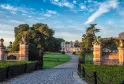 Entrée du Château Giscours avec portail en briques ornementales, allée bordée d'arbres, pelouses entretenues et château rose en arrière-plan sous un ciel bleu