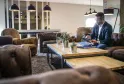 Un homme d'affaires en blazer bleu marine travaille dans le salon moderne de l'hôtel Pullman Bordeaux Lac, assis à une table en bois avec son ordinateur portable et des documents, entouré de fauteuils en cuir et d'éclairages suspendus contemporains.