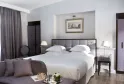 Chambre élégante de l'Hôtel de Sèze à Bordeaux avec un lit king-size à tête de lit capitonnée grise, literie blanche, accents gris-mauve, tables de nuit modernes avec lampes en laiton et un coin salon assorti