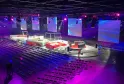 Grande arène moderne à Bordeaux avec éclairage de scène violet et bleu, estrade modulaire rouge et blanche, rangées de sièges noirs et écrans vidéo affichant des visuels dégradés