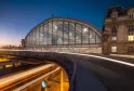 Vue nocturne de la gare de Bordeaux avec son toit en arche de verre iconique, structure métallique architecturale illuminée, immeuble haussmannien adjacent aux fenêtres éclairées, et traînées lumineuses des trains passant en dessous