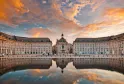 La Place de la Bourse à Bordeaux au coucher de soleil, avec ses bâtiments classiques du 18e siècle en symétrie parfaite, reflétés dans un miroir d'eau, statue équestre centrale et ciel doré dramatique.