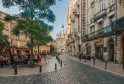Place historique de Bordeaux avec tour de château médiéval en arrière-plan, terrasse de café sous les arbres, bâtiments en pierre et esplanade piétonne par beau temps
