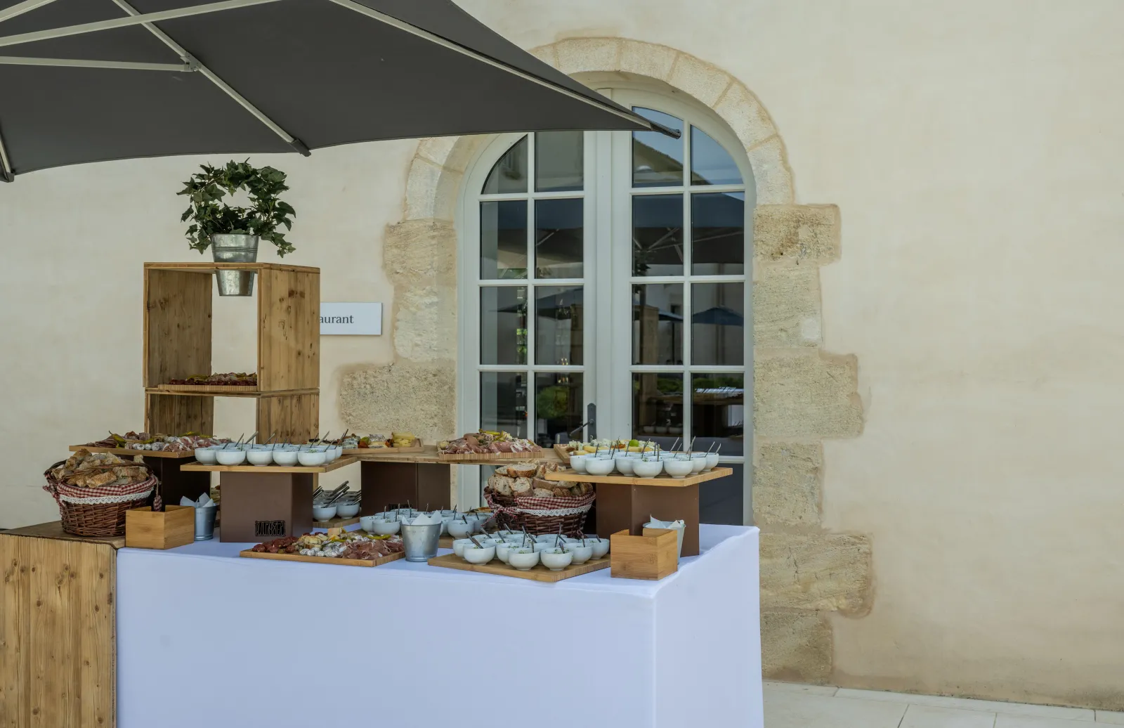 Présentoir de restauration extérieur avec bols blancs d'amuse-bouches, plateaux de charcuterie, paniers en osier et rayonnages en bois sous un parapluie noir, devant un bâtiment en pierre historique à Bordeaux.
