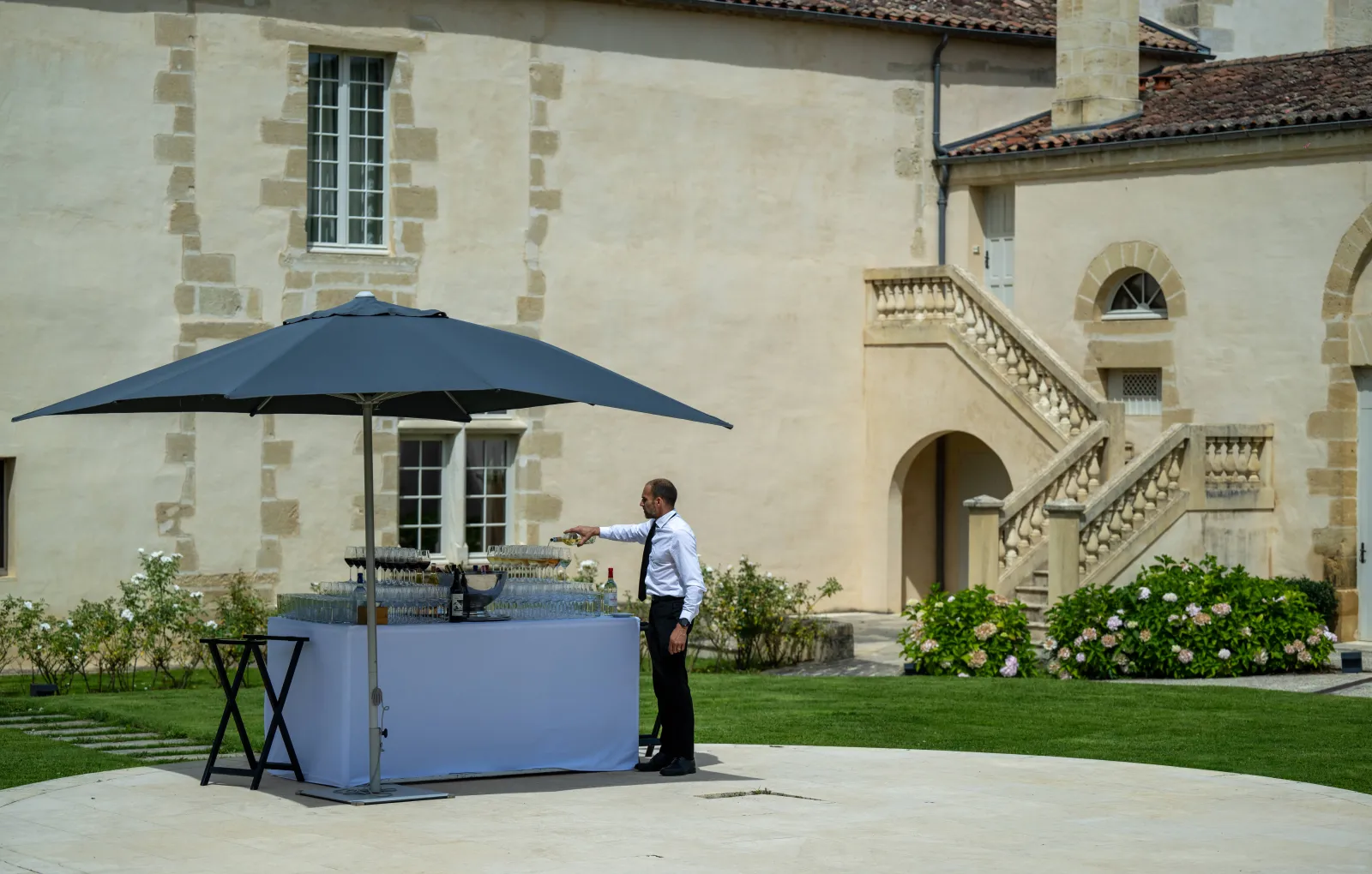 Un homme en chemise bleue se tient à un comptoir de bar mobile avec un parasol bleu foncé devant un bâtiment en pierre de style classique avec un grand escalier, entouré de fleurs blanches et d'une pelouse bien entretenue à Bordeaux.
