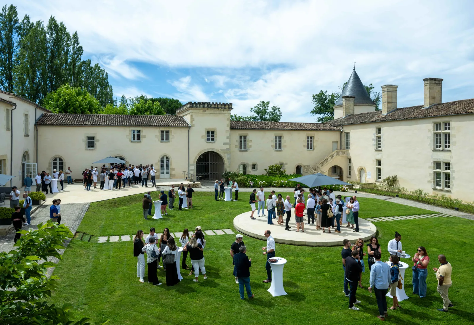 Réception de mariage en plein air dans la cour d'un château historique près de Bordeaux, avec des invités discutant sur la pelouse et des bâtiments aux murs de pierre de couleur crème présentant une architecture française traditionnelle.