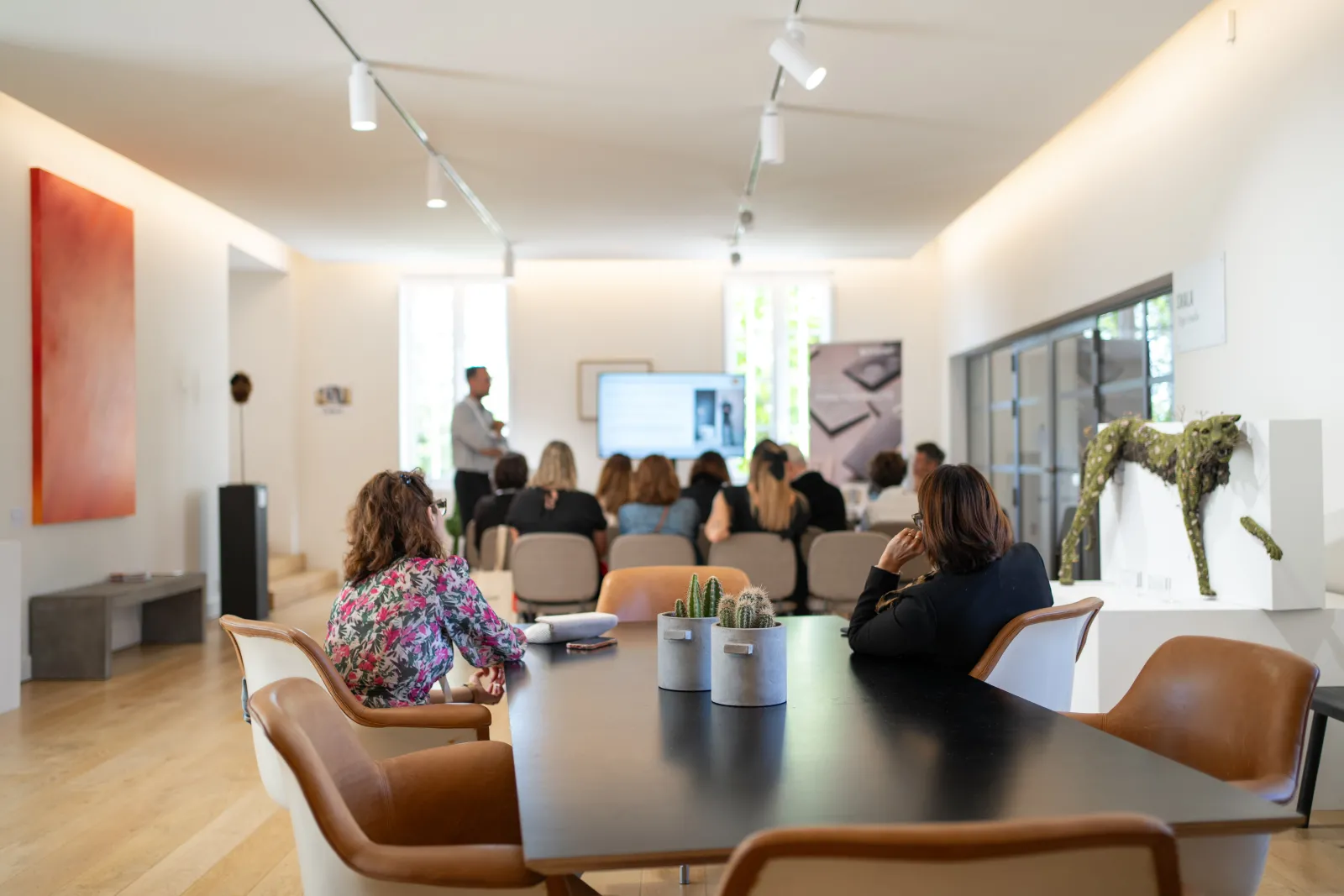 Espace de bureau moderne à Bordeaux avec une salle de conférence en arrière-plan où des employés écoutent une présentation, et un espace lounge contemporain au premier plan avec des chaises en cuir marron, des plantes en pot et une décoration épurée.