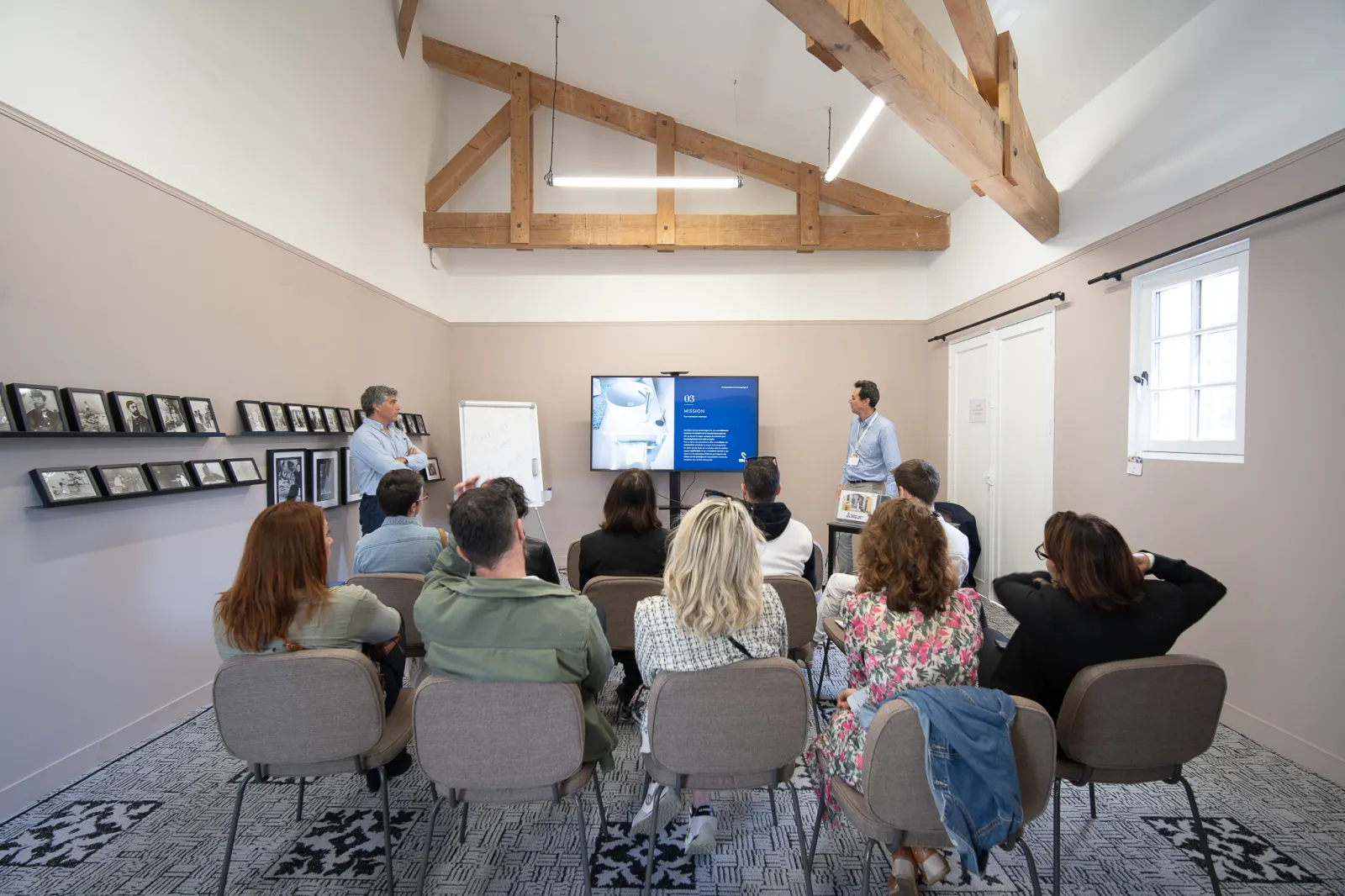 Un groupe de personnes assises dans une salle de réunion moderne à Bordeaux, face à deux présentateurs debout près d'un écran numérique bleu, avec des poutres en bois apparentes au plafond et des photographies encadrées aux murs.