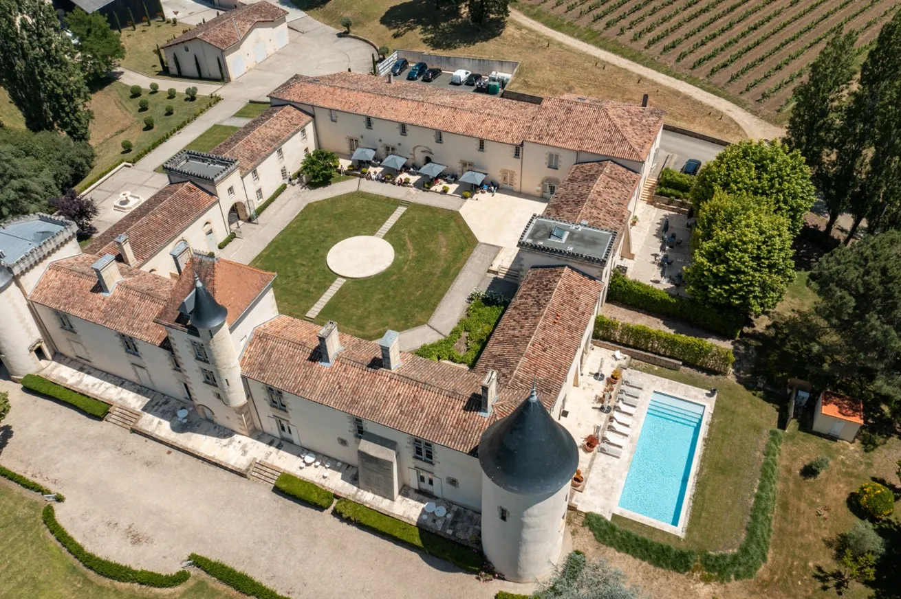 Vue aérienne d'un grand domaine de style toscan près de Bordeaux avec une villa principale aux toits en terre cuite, cour circulaire avec pelouse soignée, piscine, jardins environnants et vignobles en arrière-plan.